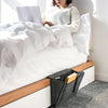 A woman is sitting on a white bed with white linens, next to a grey felt storage organizer.