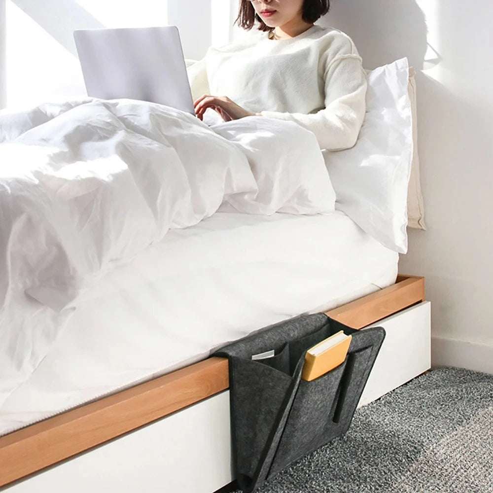 A woman is sitting on a white bed with white linens, next to a grey felt storage organizer.