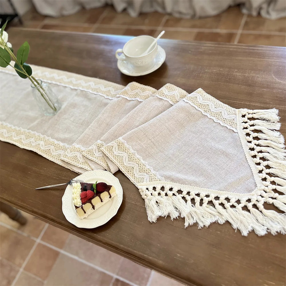 A beige crochet macrame lace table runner is displayed on a wooden table with a small cup and a piece of cake.