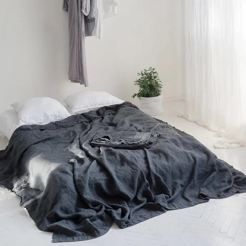 A dark gray linen flat sheet is draped on a bed, with white pillows and a potted plant in the background.