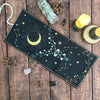 A dark fabric pouch decorated with a celestial theme featuring a crescent moon, stars, and floral elements, resting on a wood