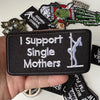 A hand holds a black iron-on patch that reads I support Single Mothers, surrounded by various other slogan patches.