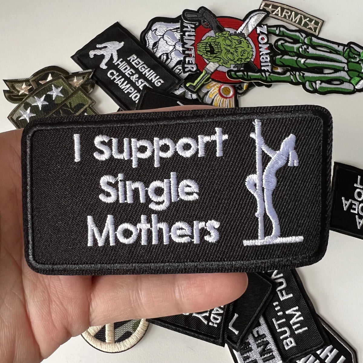 A hand holds a black iron-on patch that reads I support Single Mothers, surrounded by various other slogan patches.
