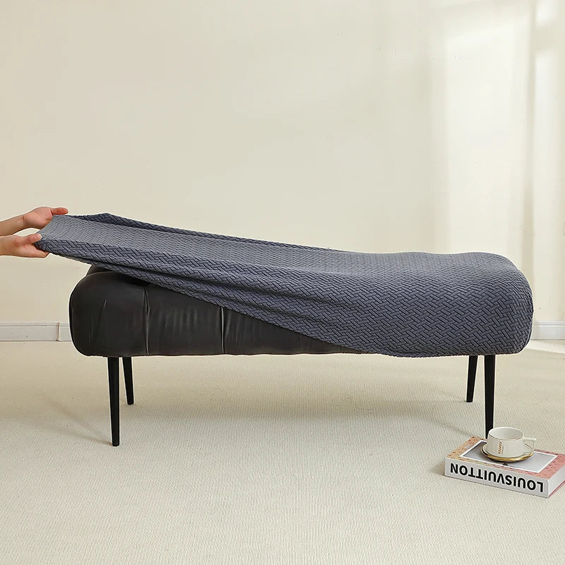 A dark gray elastic jacquard bench cover is shown on a black upholstered bench in a bright room.