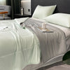 A light green reversible cooling quilt is displayed on a bed with gray bedding and pillows.