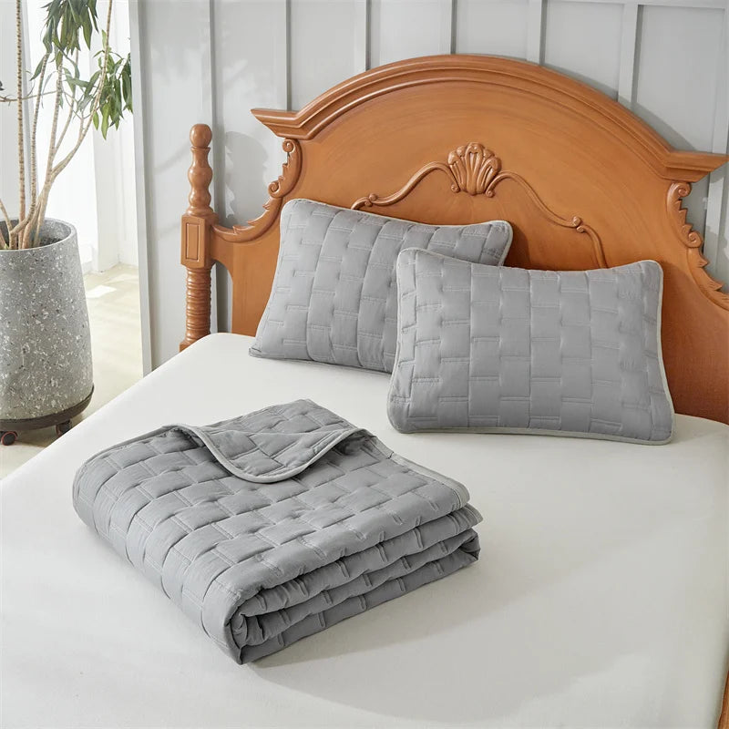 A set of light gray quilted bedding includes pillows and a folded blanket on a white bed with a wooden headboard.