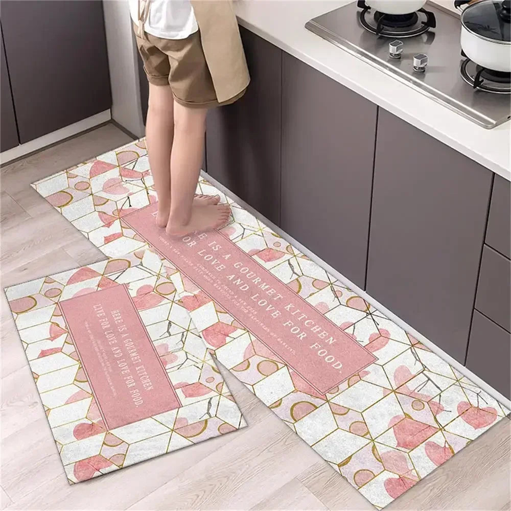 A set of two pink and white diamond print non-slip kitchen rugs placed on a light wood floor next to dark cabinets.