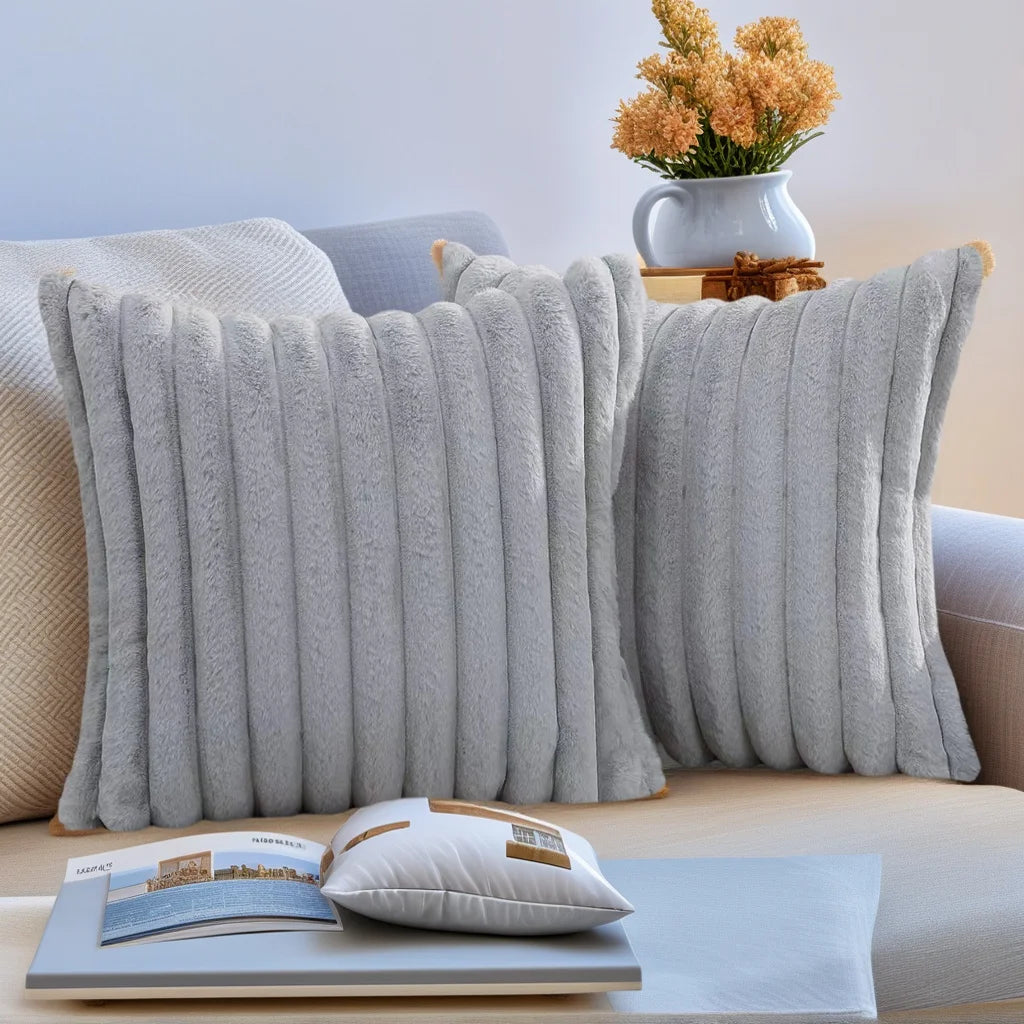 Two gray plush striped decorative pillowcases resting on a light-colored sofa with a book and a vase of flowers.