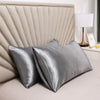 Silver satin silk pillow covers resting on a bed with a padded headboard and a lamp.