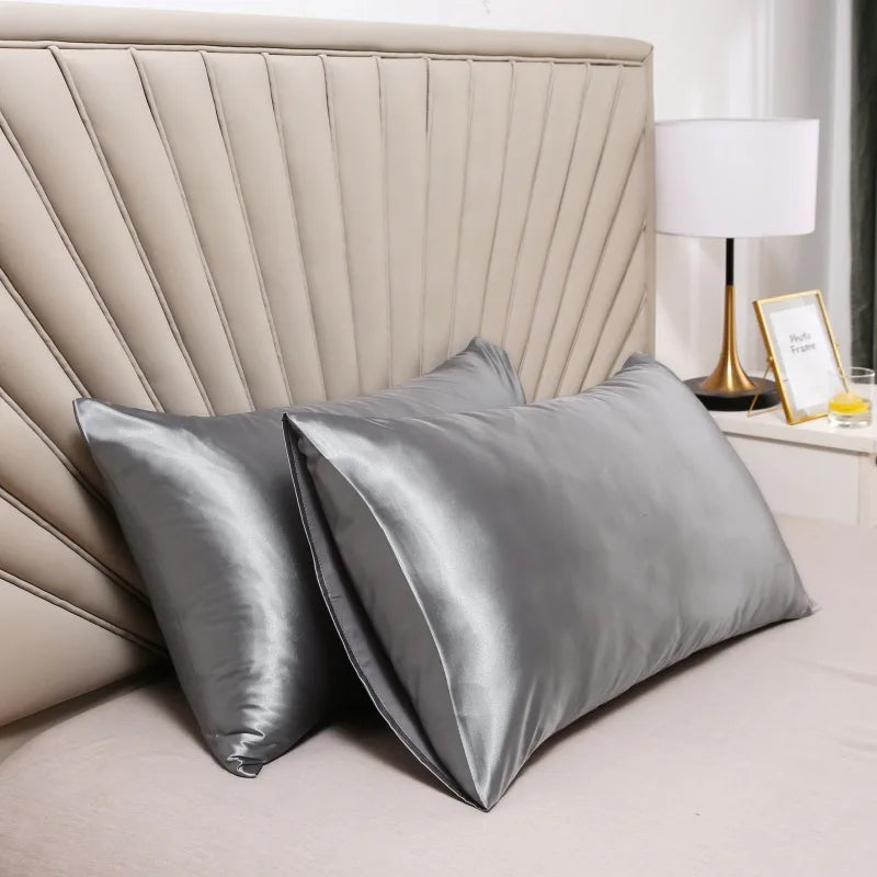 Silver satin silk pillow covers resting on a bed with a padded headboard and a lamp.