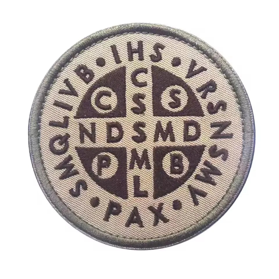 A circular embroidered patch featuring various letters and symbols in dark brown on a light background.