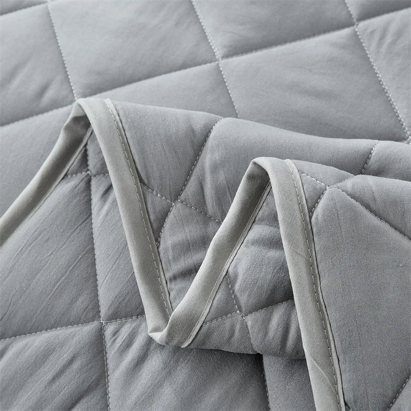 A light gray quilted bedspread set folded and draped, showing the diamond stitching pattern.