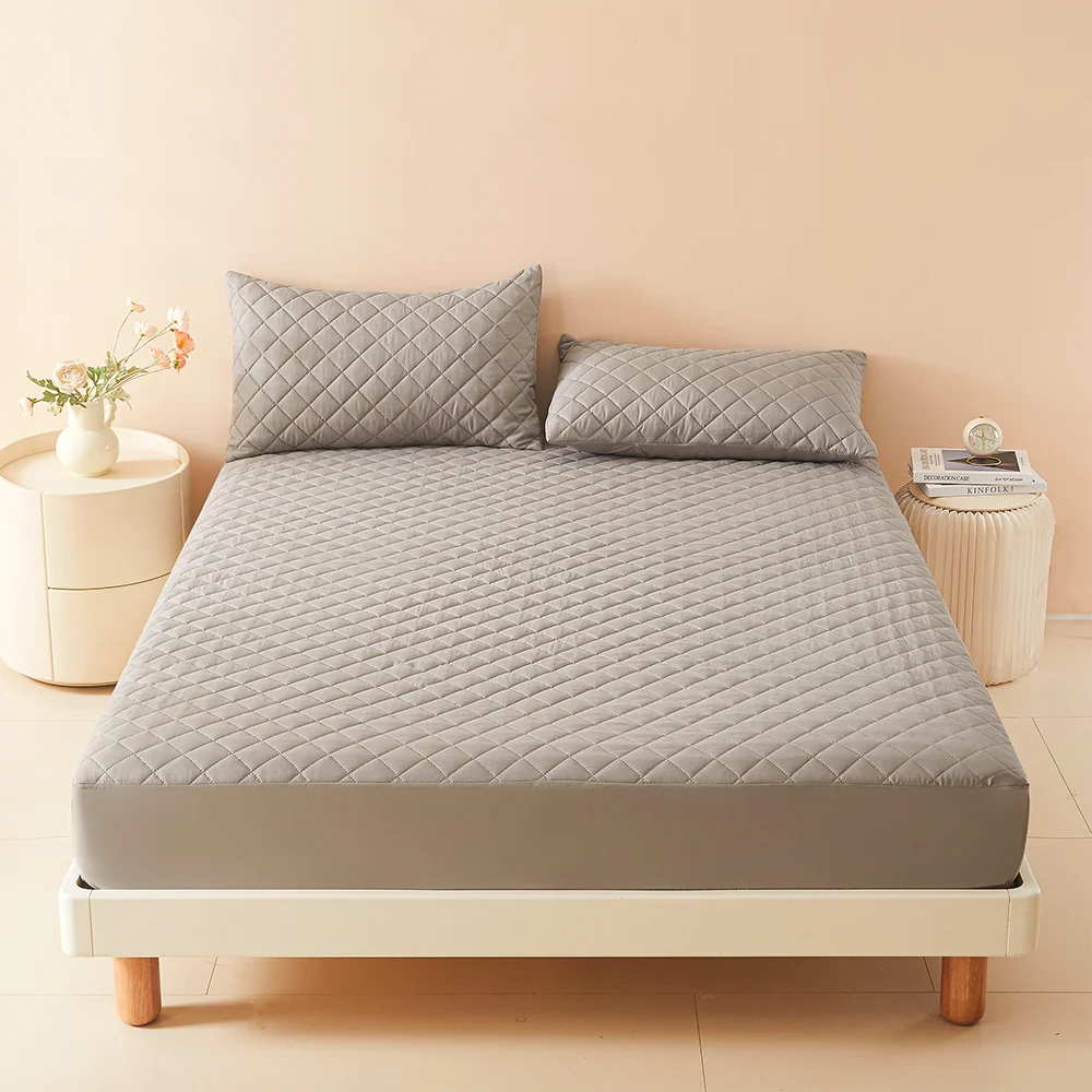 A thick gray waterproof mattress protector cover lying on a white bed frame with decorative pillows and a small vase.