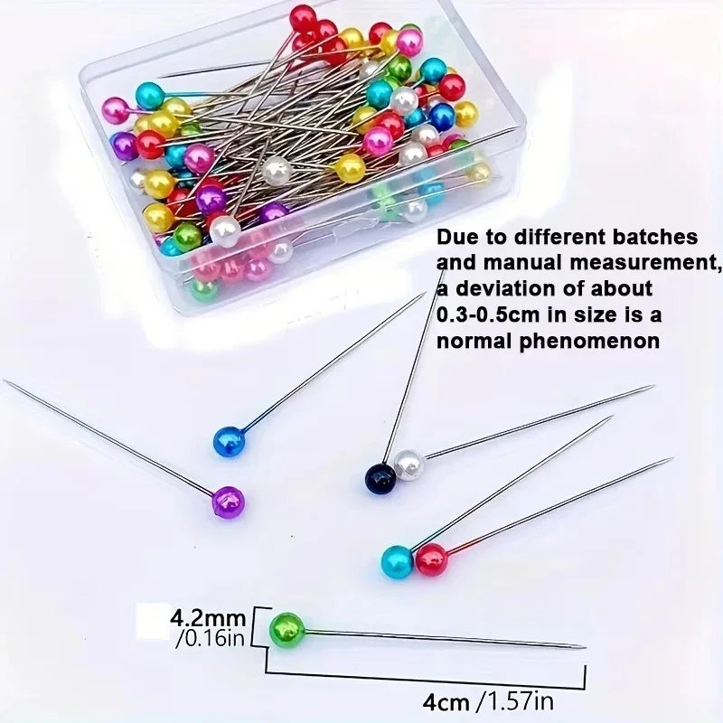 A clear container holds an assortment of colorful decorative sewing and pinning needles with varying sizes.