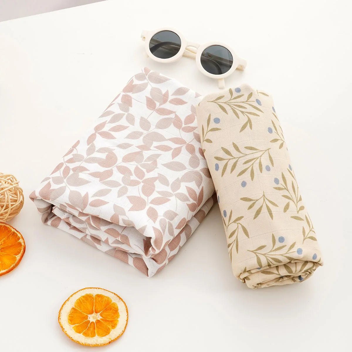 A rolled muslin swaddle blanket in beige and green floral print next to sunglasses and sliced orange.
