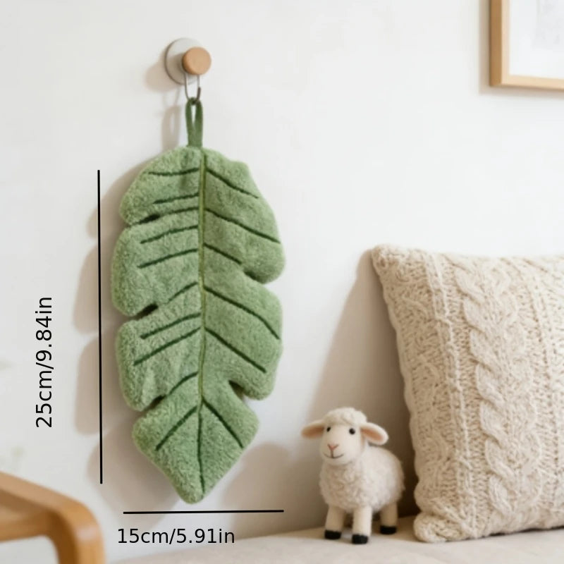 A decorative banana leaf hanging hand towel in a soft green color is displayed on a white wall next to a cream-colored knitte