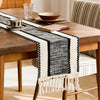 A rustic wooden table is set with a black and white macrame tassel table runner on a linen tablecloth.