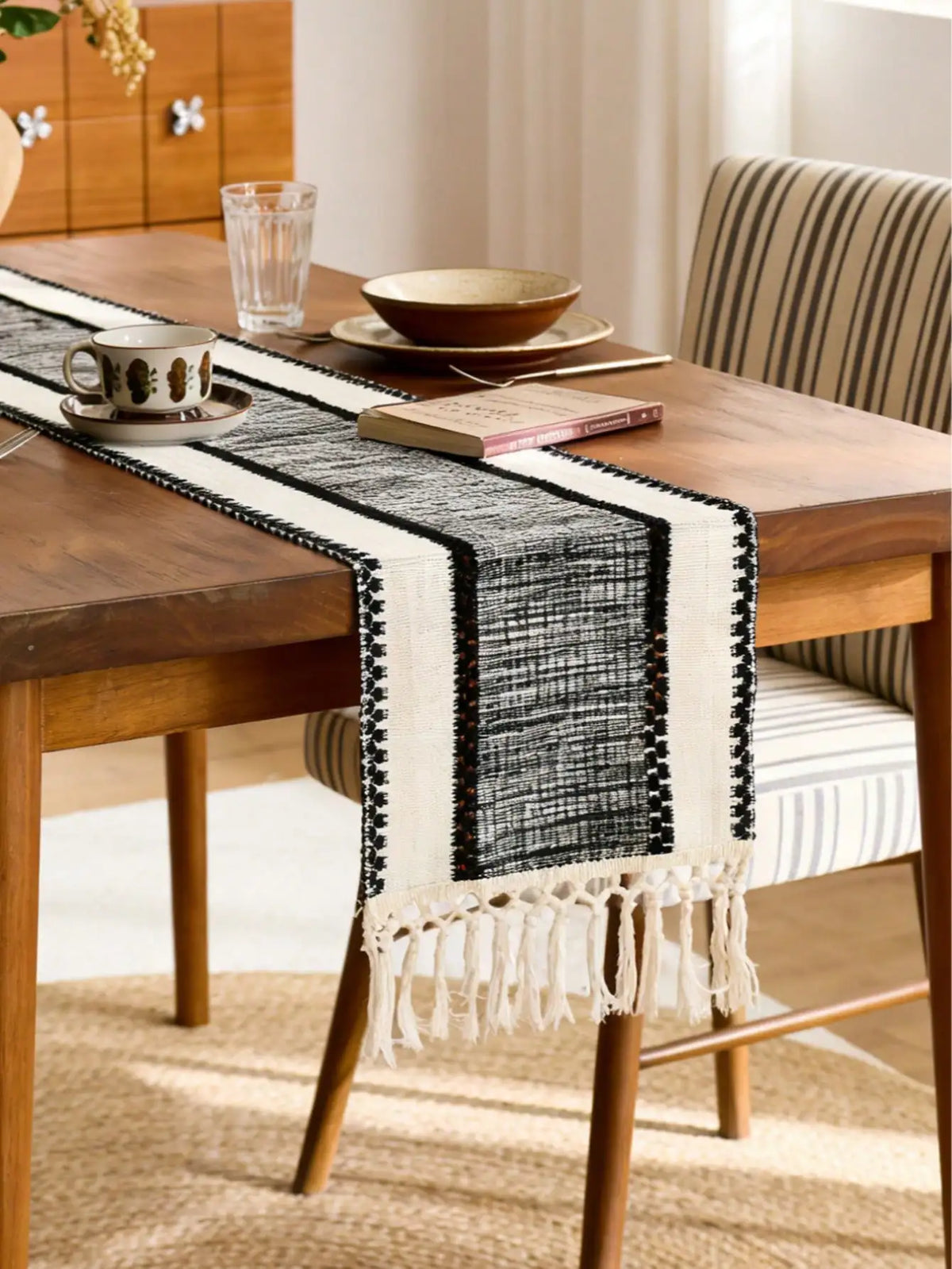 A rustic wooden table is set with a black and white macrame tassel table runner on a linen tablecloth.