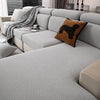 A light gray stretchy polar fleece sofa cover with a dark brown decorative pillow and a gray throw blanket on a modern sofa.