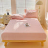 A pink and white checkered flannel fitted sheet is laid out on a wooden bed with natural light filtering through blinds.