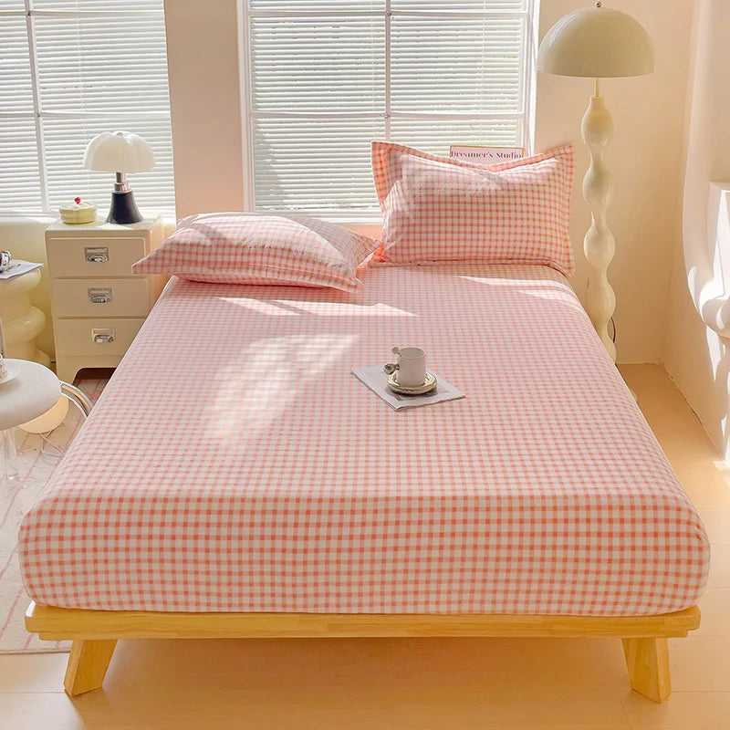 A pink and white checkered flannel fitted sheet is laid out on a wooden bed with natural light filtering through blinds.