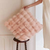 A fluffy, light pink cushion cover is being held by a hand against a white wall.