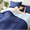 A woman is relaxing under a blue quilted blanket on a bed, with a light blue throw draped over the bedding.