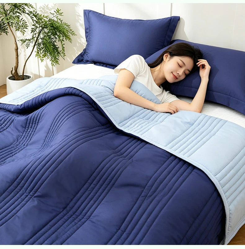A woman is relaxing under a blue quilted blanket on a bed, with a light blue throw draped over the bedding.