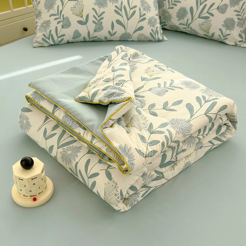 A folded Summer Gauze Cotton quilt with a light floral pattern rests on a light blue surface.