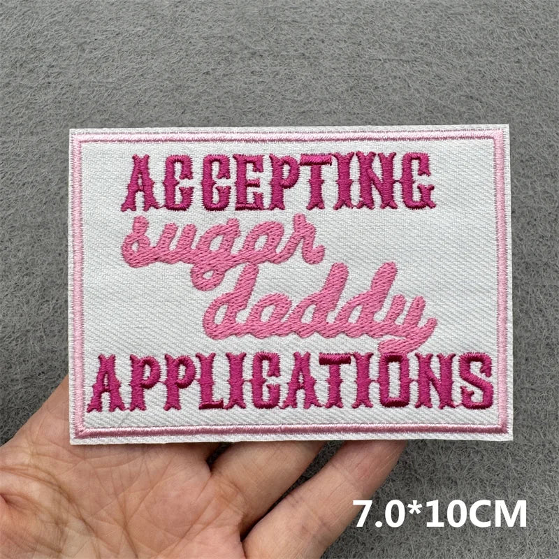 Pink embroidered patch with the text ACCEPTING SUGAR DADDY APPLICATIONS on a white fabric background