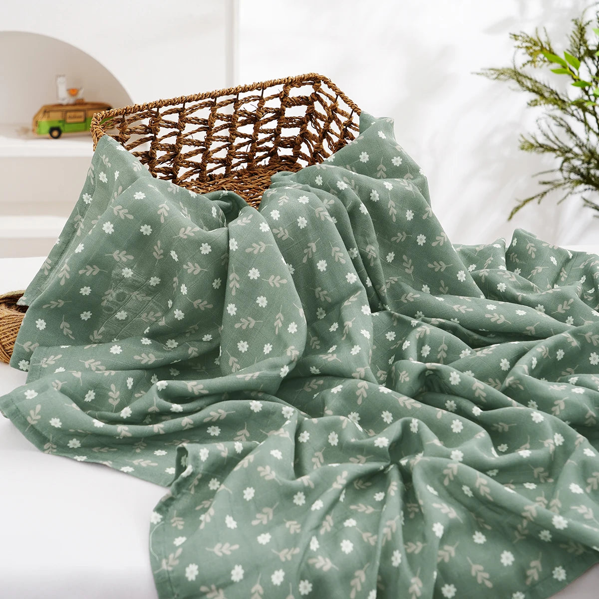 A draped sage green muslin swaddle blanket with a subtle floral pattern is displayed on a white surface.
