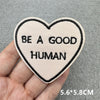 A light pink heart-shaped embroidered patch with the text BE A GOOD HUMAN is held in a hand.