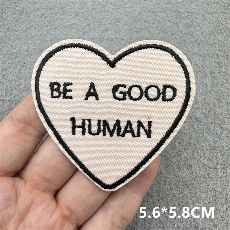 A light pink heart-shaped embroidered patch with the text BE A GOOD HUMAN is held in a hand.