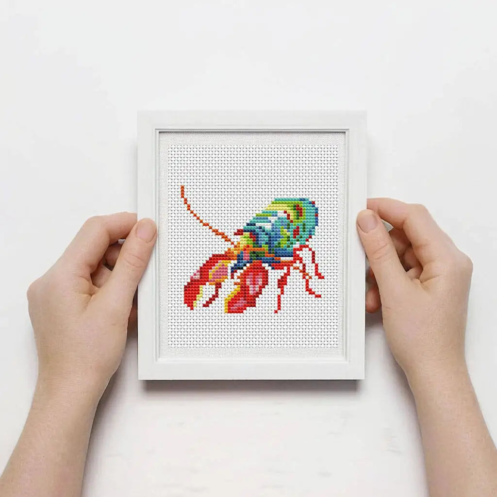 A hand holds a white framed cross stitch kit featuring a colorful fish design on a white grid.