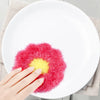 A hand places a bright pink and yellow flower-shaped kitchen dish scrubber sponge on a white plate.