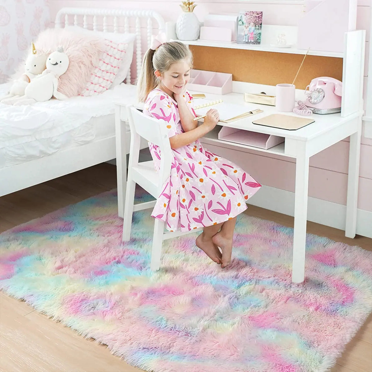 A young girl sits at a white desk in a bedroom, decorating or writing on paper, with a colorful fluffy rug beneath her.