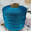 A spool of bright turquoise glitter metallic yarn for crochet and knitting resting on a white surface with pink flowers.