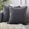 A dark gray velvet decorative cushion cover with black tasseled trim resting on a light cream knitted blanket.