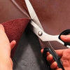 A pair of silver fabric scissors is shown cutting through brown leather material.