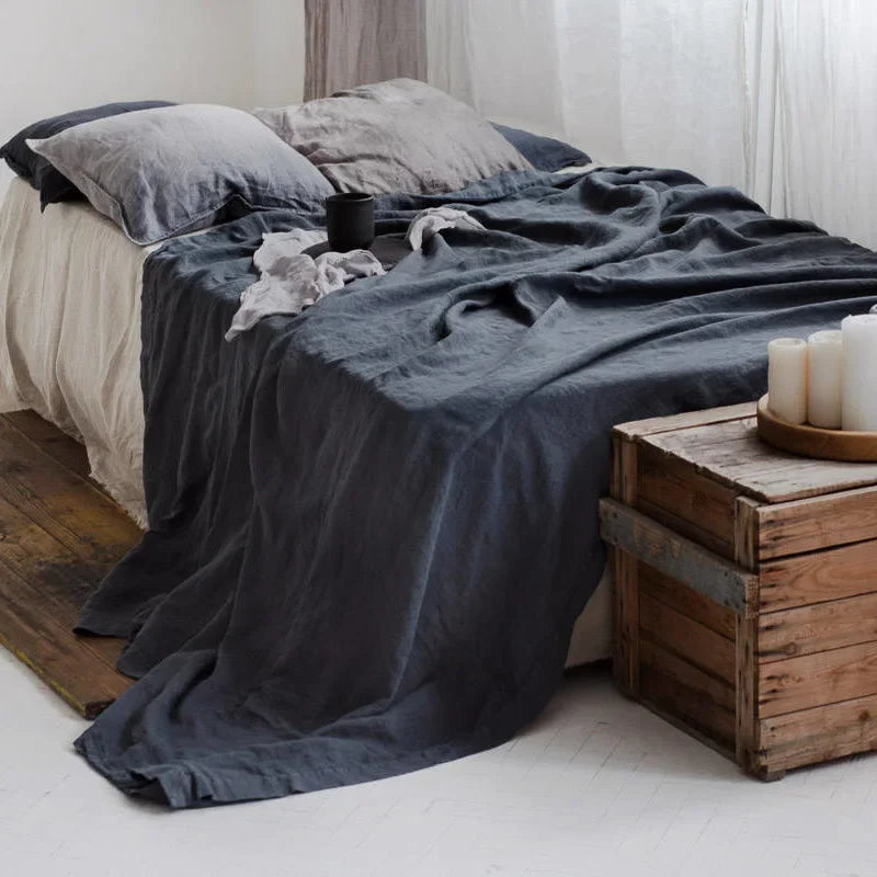 A dark gray linen flat sheet draped over a bed with light gray pillows and wooden boxes.