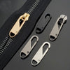 A set of brass and silver zipper pullers and a black zipper tape are displayed on a dark surface.
