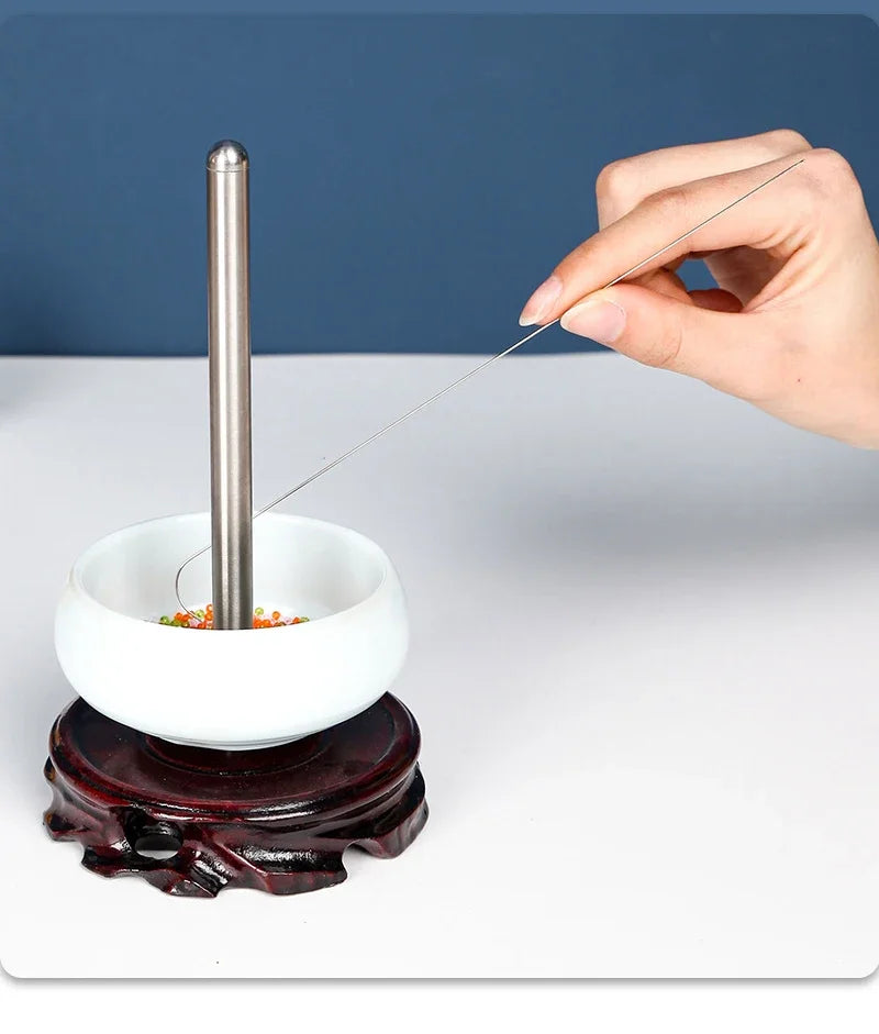 A hand uses a thin metal needle to thread a bead onto a stainless steel bead needle set for crafting.