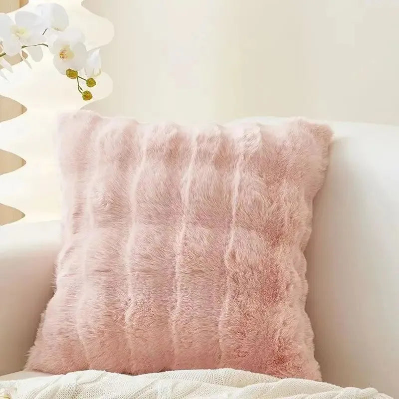 A fluffy, light pink faux fur throw pillow resting on a white surface next to a white plant.