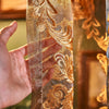 A hand holds a sheer curtain fabric featuring intricate gold and bronze floral embroidery.