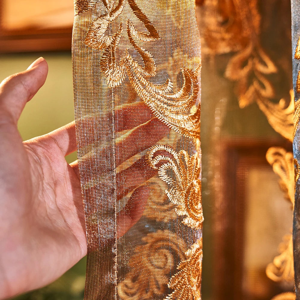 A hand holds a sheer curtain fabric featuring intricate gold and bronze floral embroidery.