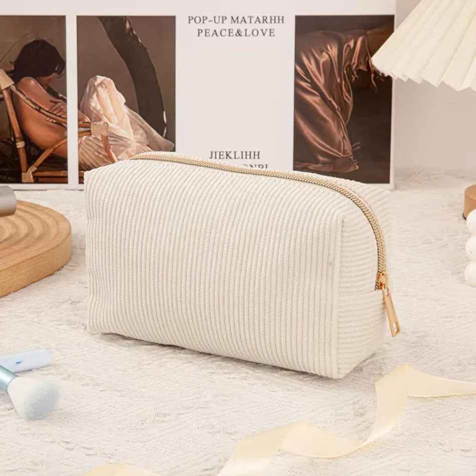A cream-colored, ribbed cosmetic storage pouch with a gold zipper lies on a white fluffy rug.