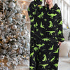 A woman models a black sherpa fleece oversized blanket hoodie with green dinosaur print in a bright indoor setting.