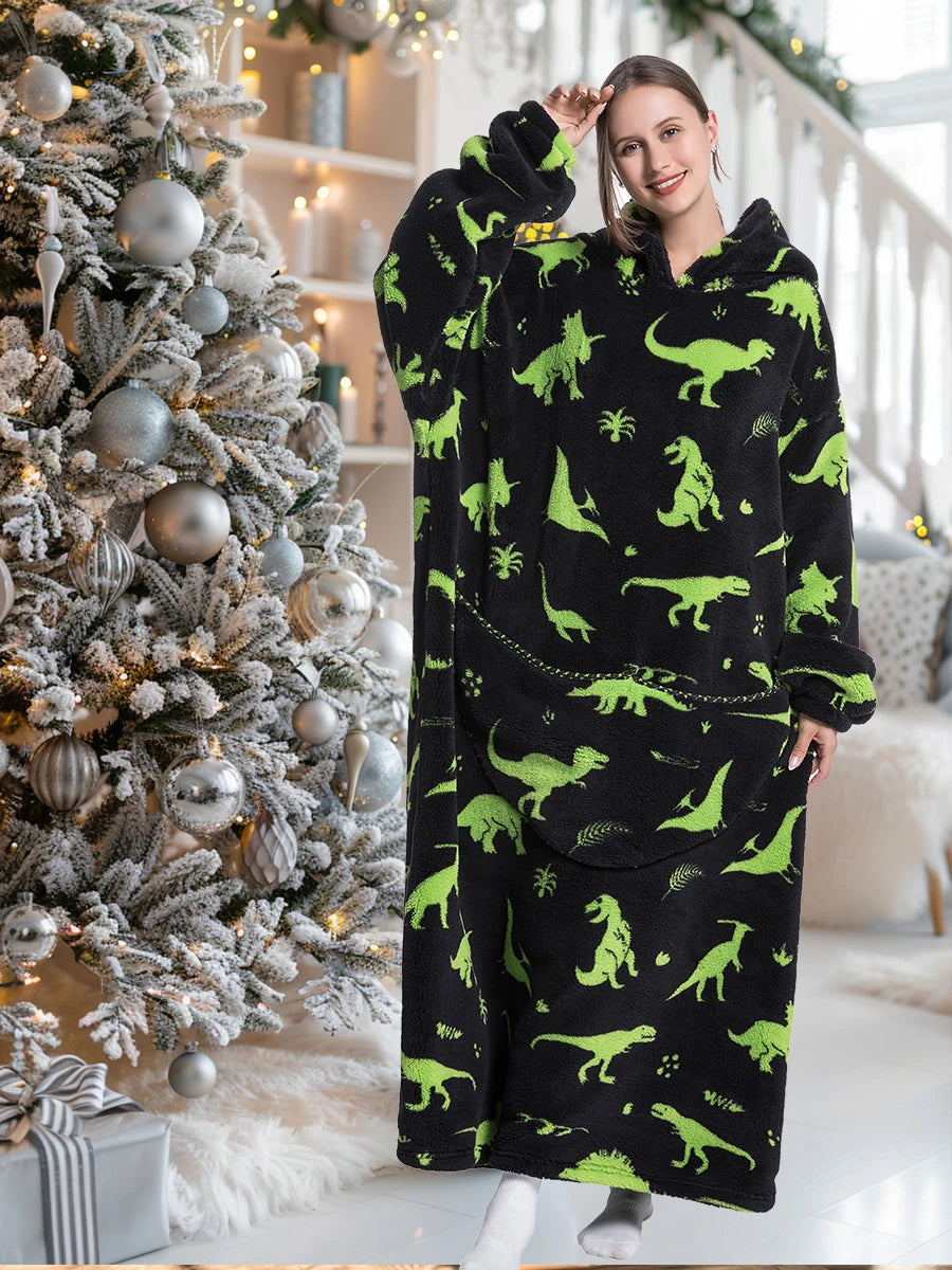 A woman models a black sherpa fleece oversized blanket hoodie with green dinosaur print in a bright indoor setting.