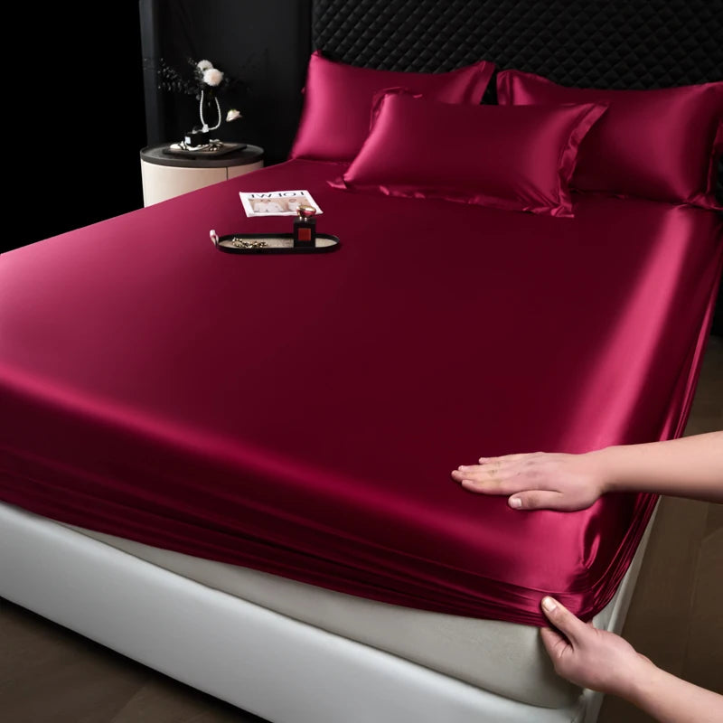 A deep burgundy satin fitted sheet is laid out on a bed, with a hand touching the fabric.