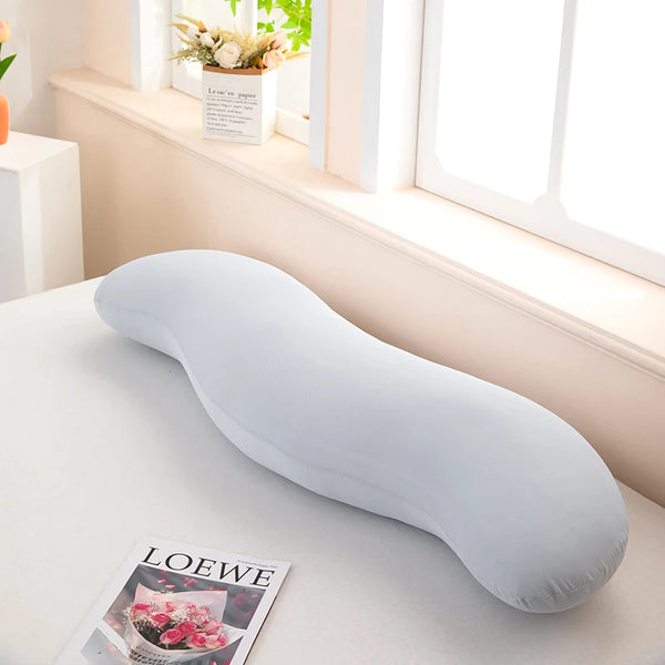 A light gray ergonomic double wave curve pillow resting on a white surface next to a window sill.